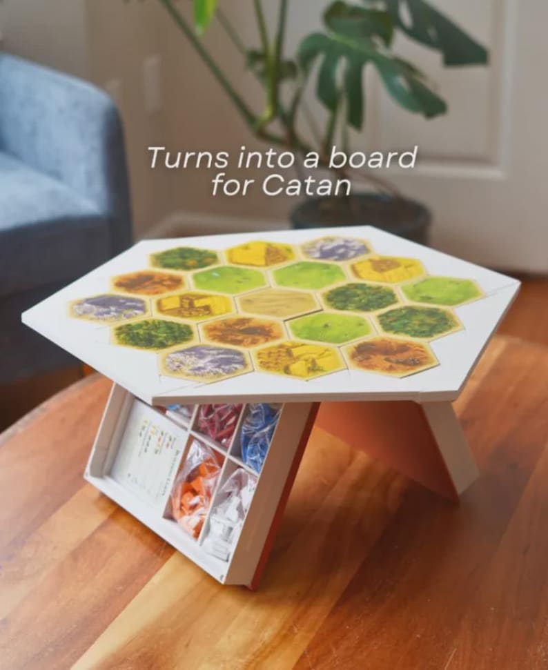 May include: A white hexagonal board game table with a colorful Catan game board design. The table has a built-in storage compartment with game pieces. The text "Turns into a board for Catan" is visible on the image.