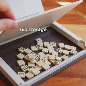 May include: A white and gray tile storage box with a hinged lid, open to reveal a collection of off-white letter tiles. The text "Tile storage" is visible on the box. The tiles are scattered on a dark brown surface.