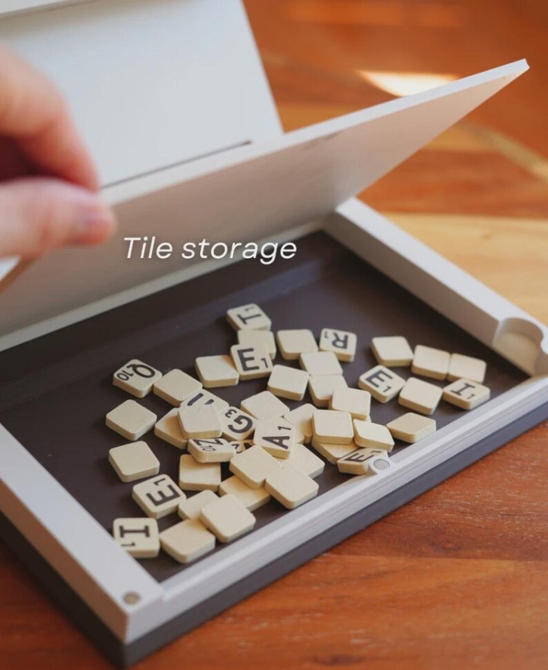 May include: A white and gray tile storage box with a hinged lid, open to reveal a collection of off-white letter tiles. The text "Tile storage" is visible on the box. The tiles are scattered on a dark brown surface.
