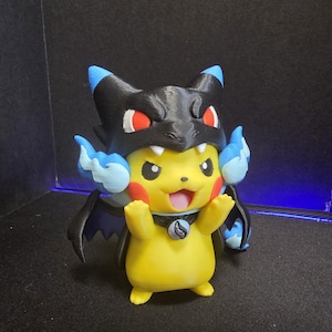 May include: A yellow Pikachu figurine wearing a black Charizard hood with red eyes and blue flames. The character has red cheeks, a wide smile, and a small circular pendant around its neck. It features black wings and a blue-tipped tail.