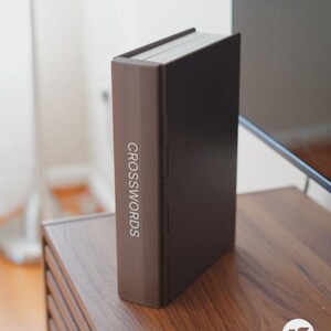 May include: A brown book-shaped box with the word "CROSSWORDS" printed vertically on the spine. The box is closed and sits upright on a wooden surface. The box has a dark brown front and back cover.