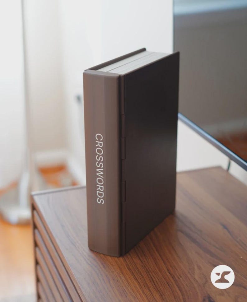 May include: A brown book-shaped box with the word "CROSSWORDS" printed vertically on the spine. The box is closed and sits upright on a wooden surface. The box has a dark brown front and back cover.