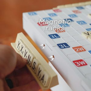 May include: A close-up of a Scrabble board with magnetic racks. The board is white with blue, red, and orange squares. The magnetic racks are beige, and the tiles have black letters and numbers. The text "Magnetic racks" is visible.