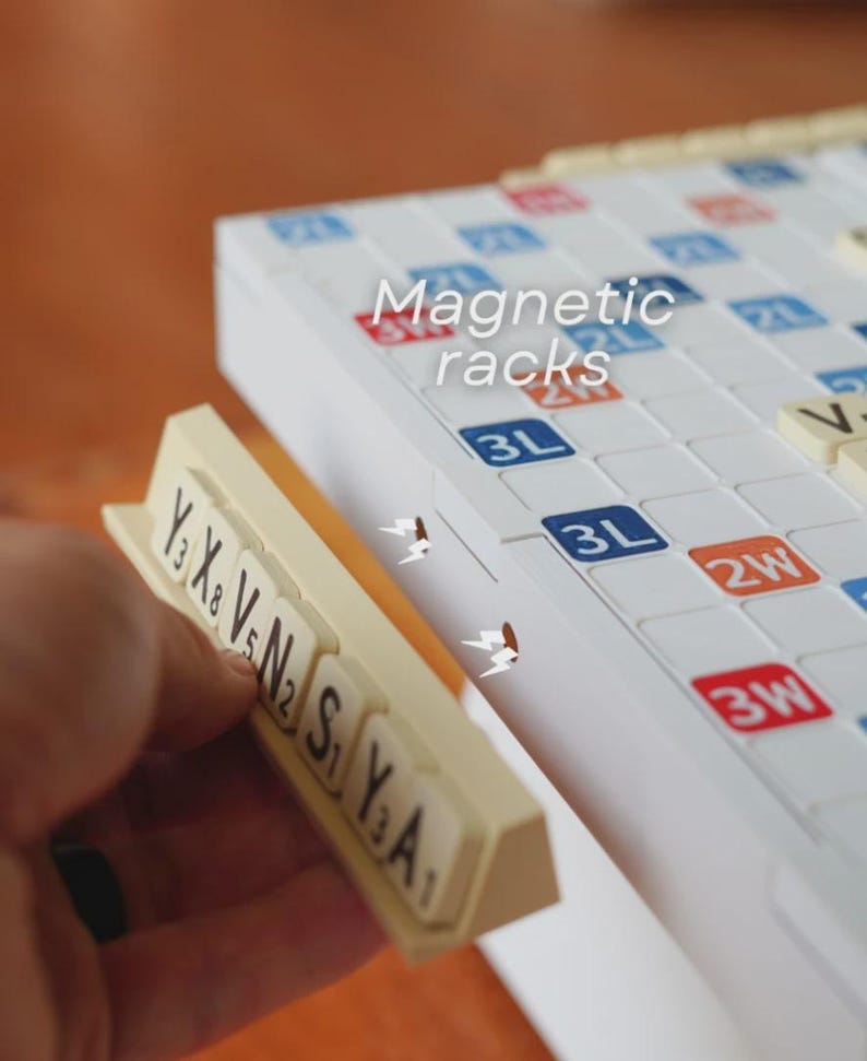 May include: A close-up of a Scrabble board with magnetic racks. The board is white with blue, red, and orange squares. The magnetic racks are beige, and the tiles have black letters and numbers. The text "Magnetic racks" is visible.