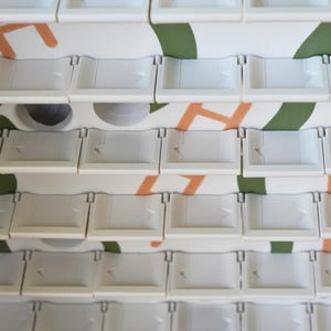 May include: Close-up of a stack of white plastic trays with a small arrow design. The trays are arranged in rows and columns. The background features a white surface with orange and green abstract shapes.
