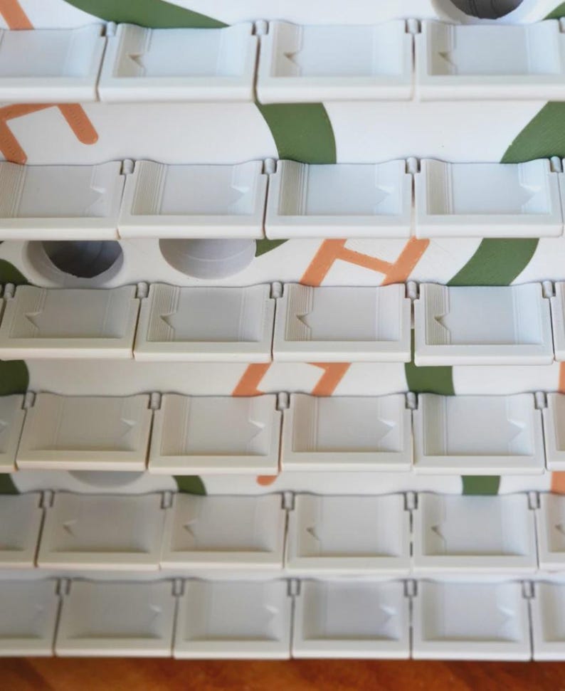 May include: Close-up of a stack of white plastic trays with a small arrow design. The trays are arranged in rows and columns. The background features a white surface with orange and green abstract shapes.