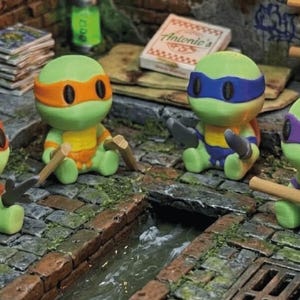 Op de afbeelding: Vier kleine, groene schildpadfiguurtjes in cartoonstijl die op een stenen ondergrond zitten. Elke schildpad draagt een masker in een andere kleur: oranje, blauw of paars. Ze houden miniatuurwapens vast, waaronder nunchucks, katana's en een bo-staf.