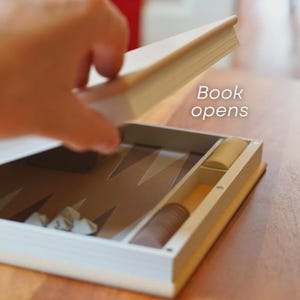 Puede incluir: Un juego de backgammon dise&ntilde;ado como un libro, con el texto "Book opens". El juego est&aacute; abierto, revelando el tablero y las piezas. El tablero es marr&oacute;n y beige, con piezas blancas y marrones.