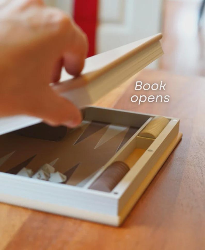 Puede incluir: Un juego de backgammon dise&ntilde;ado como un libro, con el texto "Book opens". El juego est&aacute; abierto, revelando el tablero y las piezas. El tablero es marr&oacute;n y beige, con piezas blancas y marrones.