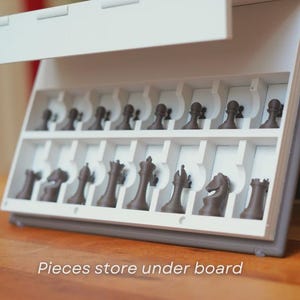 May include: A white and gray chess set with brown chess pieces. The pieces are stored in a compartment under the board. The text "Pieces store under board" is visible on the bottom of the image.