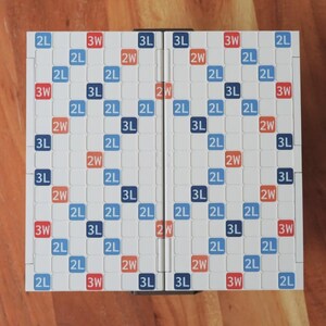 May include: A Scrabble board game, folded open, featuring a grid of white squares with blue, red, and orange text. The board is set on a wooden surface. The game board is designed for word-building.