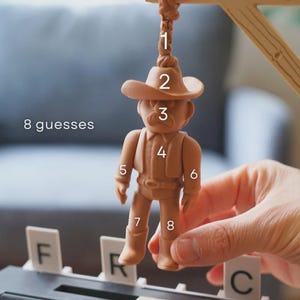 May include: A tan cowboy figurine suspended from a wooden gallows, part of a game. The figurine wears a hat, shirt, and boots. The text "8 guesses" is visible. The letters F, R, and C are also present.