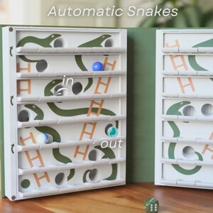 May include: A white wooden board game with green snake illustrations and the text "Automatic Snakes." The game features a series of holes, ladders, and a dice. Several colored marbles are visible inside the game.