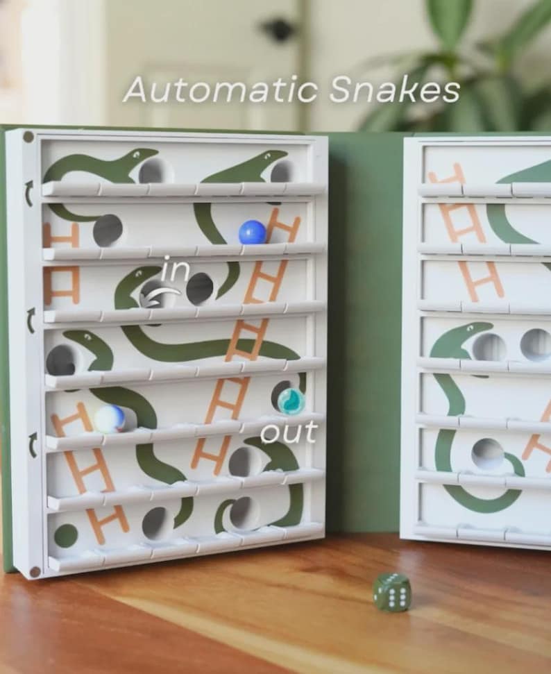 May include: A white wooden board game with green snake illustrations and the text "Automatic Snakes." The game features a series of holes, ladders, and a dice. Several colored marbles are visible inside the game.