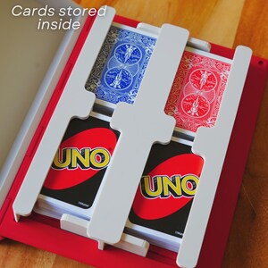 May include: A red card holder with white dividers, holding decks of UNO and playing cards. The text "Cards stored inside" is visible. The UNO cards are red and yellow. The playing cards have blue and red backs.