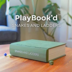 May include: A green "PlayBook'd" Snakes and Ladders game set on a wooden table. The game is designed to look like a book. Two small game pieces and a die are next to the game. The background includes a plant and a gray chair.