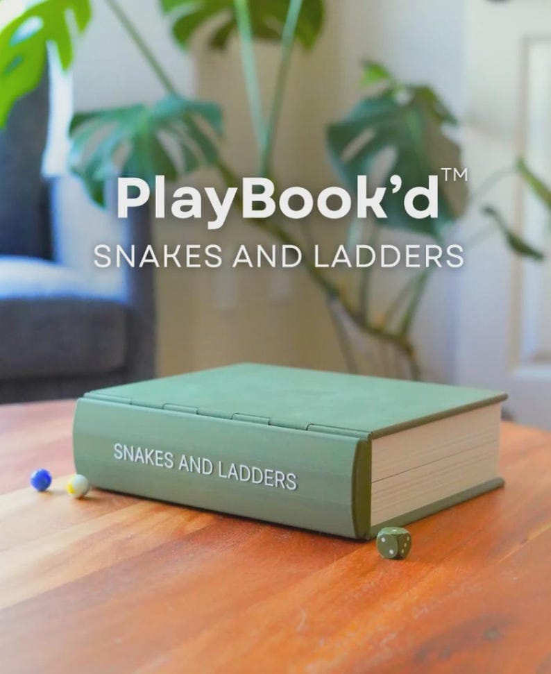May include: A green "PlayBook'd" Snakes and Ladders game set on a wooden table. The game is designed to look like a book. Two small game pieces and a die are next to the game. The background includes a plant and a gray chair.