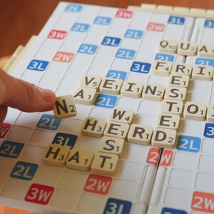 May include: A Scrabble board with beige letter tiles spelling out words. A hand is placing the letter "N" on the board. The board has a white grid with colored squares, including blue, red, and orange.