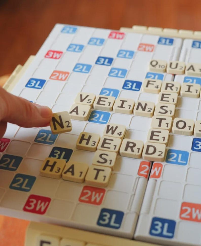 May include: A Scrabble board with beige letter tiles spelling out words. A hand is placing the letter "N" on the board. The board has a white grid with colored squares, including blue, red, and orange.