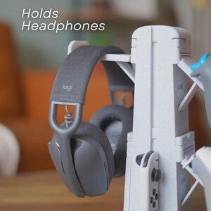 Może przedstawiać: Szare słuchawki spoczywają na białym stojaku na słuchawki. Stojak mieści r&oacute;wnież biały kontroler do gier. Tekst "Holds Headphones" jest widoczny. Słuchawki mają markę "logi" na pałąku.