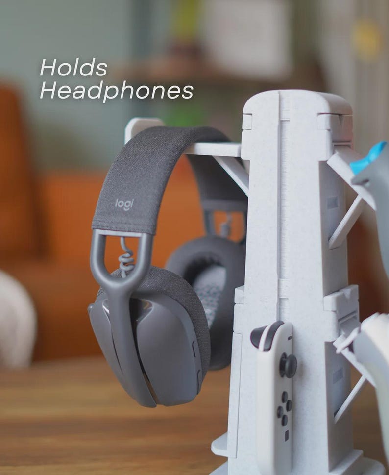 Może przedstawiać: Szare słuchawki spoczywają na białym stojaku na słuchawki. Stojak mieści r&oacute;wnież biały kontroler do gier. Tekst "Holds Headphones" jest widoczny. Słuchawki mają markę "logi" na pałąku.