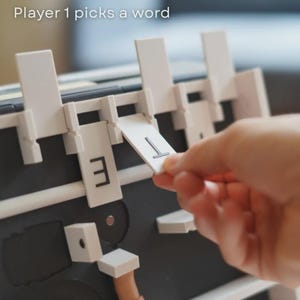 May include: A board game with white and black elements is shown. The game has a letter selection mechanism. The text "Player 1 picks a word" is visible. A hand is placing a letter card with the letter "T".