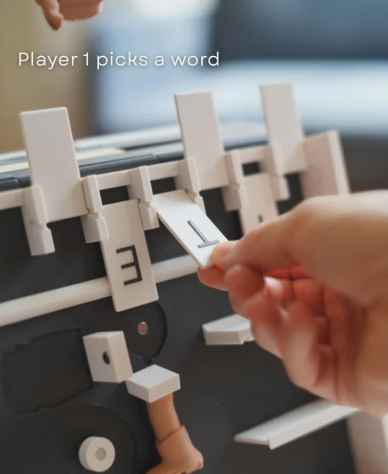 May include: A board game with white and black elements is shown. The game has a letter selection mechanism. The text "Player 1 picks a word" is visible. A hand is placing a letter card with the letter "T".