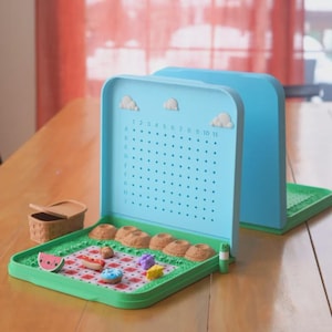 Könnte beinhalten: Ein blau-grünes magnetisches Brettspiel zum Thema Picknick. Das Spiel zeigt einen Picknickkorb, Lebensmittel und eine karierte Picknickdecke. Das Brett hat ein Raster mit Zahlen und Buchstaben sowie dekorative Wolken.