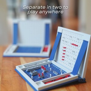 Op de afbeelding: Een klassiek Zeeslag-spel in een blauw en witte behuizing. Het spelbord is open en onthult een blauw raster met miniatuurschepen en pinnen. De tekst "Separate in two to play anywhere" is zichtbaar.
