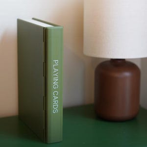 May include: A green playing card case stands upright on a green surface. The words "PLAYING CARDS" are printed vertically on the spine in white. A brown lamp with a white lampshade is in the background.