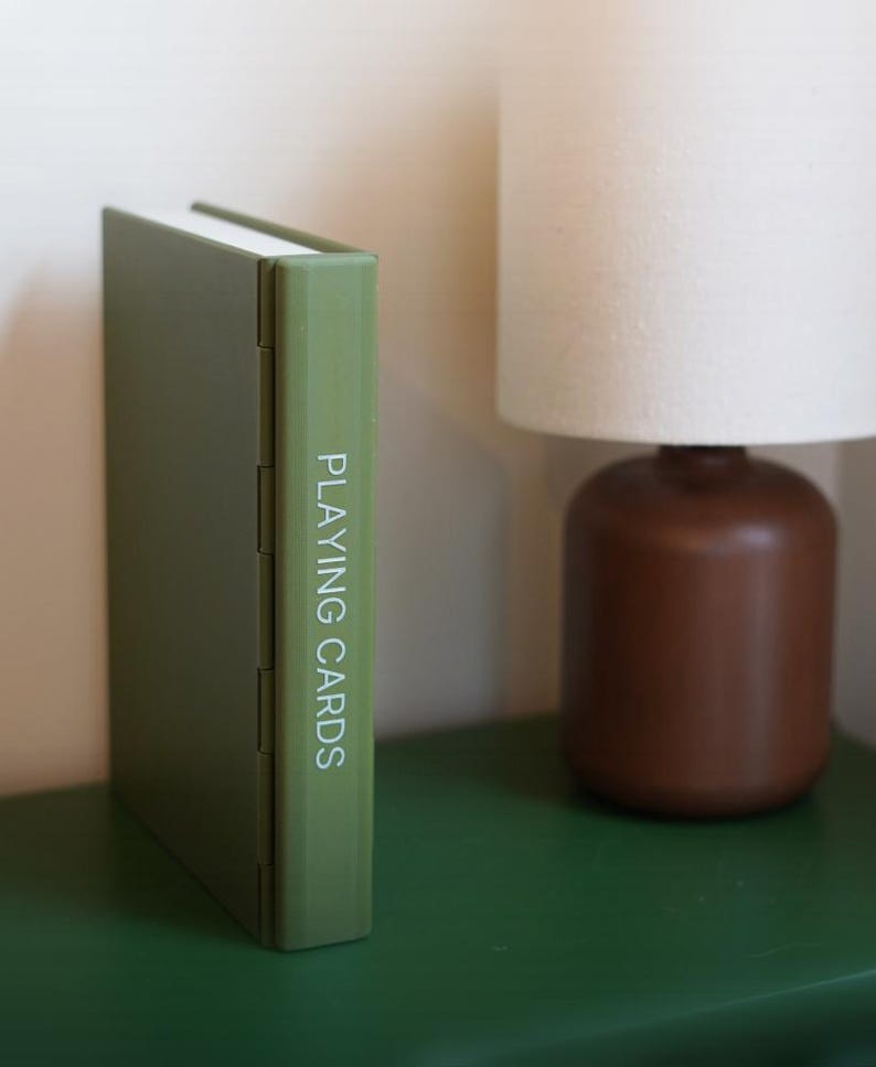 May include: A green playing card case stands upright on a green surface. The words "PLAYING CARDS" are printed vertically on the spine in white. A brown lamp with a white lampshade is in the background.