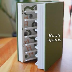 May include: A green book with white-edged pages is partially open, revealing a series of shelves with a snake design. The text "Book opens" is visible on the cover. The book is on a wooden surface.
