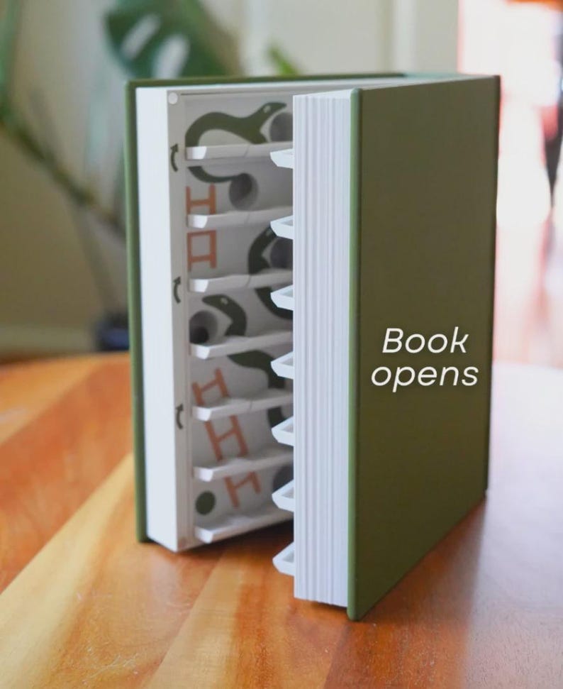May include: A green book with white-edged pages is partially open, revealing a series of shelves with a snake design. The text "Book opens" is visible on the cover. The book is on a wooden surface.