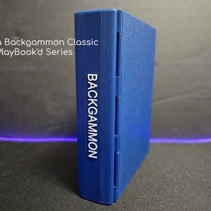May include: A blue, rectangular, foldable backgammon game case with a textured surface. The spine features the word BACKGAMMON in white text, while the upper left area displays the text Magna Backgammon Classic PlayBook'd Series.