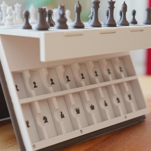 May include: A white and black portable chess set. The chess pieces are black and white. The set has a built-in storage compartment for the chess pieces. The chess pieces are arranged on the board, ready for play.