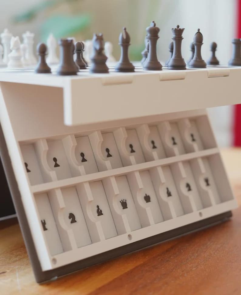 May include: A white and black portable chess set. The chess pieces are black and white. The set has a built-in storage compartment for the chess pieces. The chess pieces are arranged on the board, ready for play.