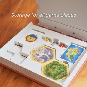 May include: Open board game box with white interior and orange trim. The box contains game pieces, including cards, dice, and hexagonal tiles depicting landscapes. Text on the box reads "Storage for all game pieces."