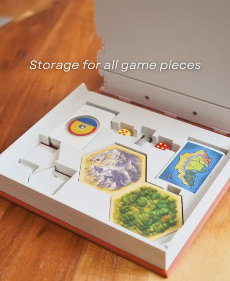 May include: Open board game box with white interior and orange trim. The box contains game pieces, including cards, dice, and hexagonal tiles depicting landscapes. Text on the box reads "Storage for all game pieces."