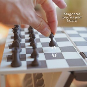 May include: A close-up of a magnetic chess set with black and white squares. The dark brown chess pieces are arranged on the board. The text "Magnetic pieces and board" is visible. A hand is placing a pawn on the board.