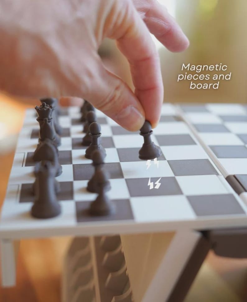 May include: A close-up of a magnetic chess set with black and white squares. The dark brown chess pieces are arranged on the board. The text "Magnetic pieces and board" is visible. A hand is placing a pawn on the board.