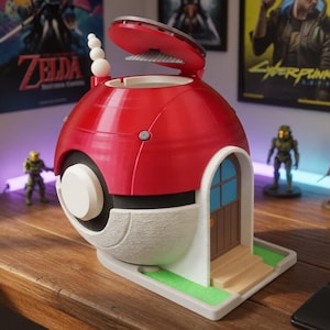 May include: A red and white spherical container with a hinged top, resembling a Poké Ball, with a small door and steps. The container is on a wooden surface, with figurines and posters in the background.