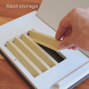 May include: A white and beige rack storage unit with a hand placing a beige divider. The unit has a rectangular frame with multiple beige dividers. The text "Rack storage" is visible in the upper left corner.