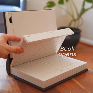 May include: An open book with a dark brown cover and white pages. The text "Book opens" is visible. The book is on a wooden surface, with a plant in the background.