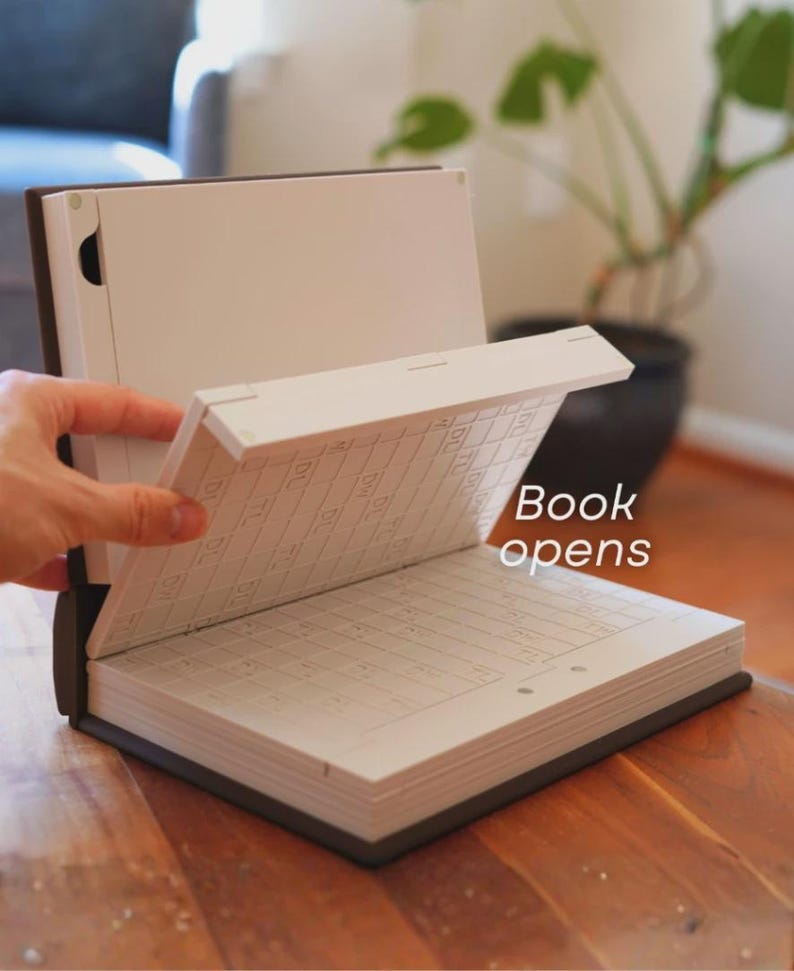 May include: An open book with a dark brown cover and white pages. The text "Book opens" is visible. The book is on a wooden surface, with a plant in the background.