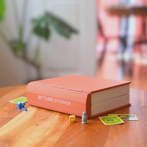 May include: A coral-colored Settlers Storage box, designed to look like a book, with the text "SETTLERS STORAGE" in white. The box is on a wooden table, with game pieces and cards scattered around it. A yellow die and a gray game piece are visible.