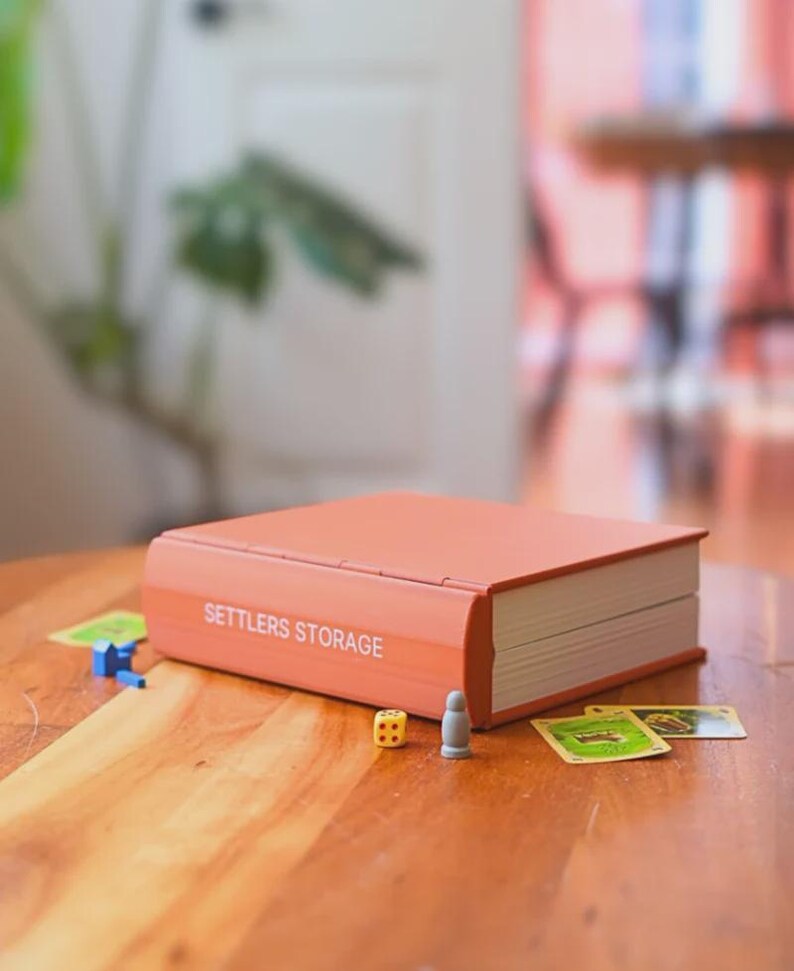 May include: A coral-colored Settlers Storage box, designed to look like a book, with the text "SETTLERS STORAGE" in white. The box is on a wooden table, with game pieces and cards scattered around it. A yellow die and a gray game piece are visible.