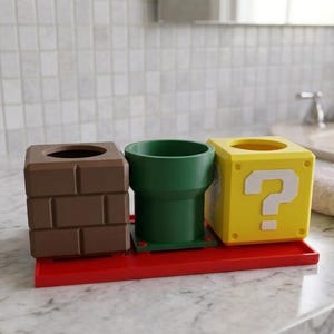 May include: A set of bathroom accessories inspired by the Super Mario Bros. video game. The set includes a brown brick-like container, a green pipe-shaped cup, and a yellow question mark block, all on a red tray.
