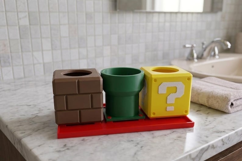 May include: A set of bathroom accessories inspired by the Super Mario Bros. video game. The set includes a brown brick-like container, a green pipe-shaped cup, and a yellow question mark block, all on a red tray.