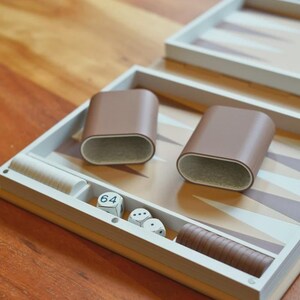 Puede incluir: Un juego de backgammon con una superficie de juego marr&oacute;n claro y beige. El juego incluye dos cubiletes de dados de cuero marr&oacute;n, dados y fichas de madera. El tablero est&aacute; abierto, listo para jugar.
