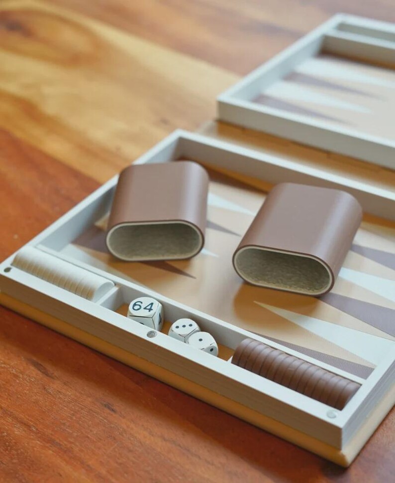 Puede incluir: Un juego de backgammon con una superficie de juego marr&oacute;n claro y beige. El juego incluye dos cubiletes de dados de cuero marr&oacute;n, dados y fichas de madera. El tablero est&aacute; abierto, listo para jugar.